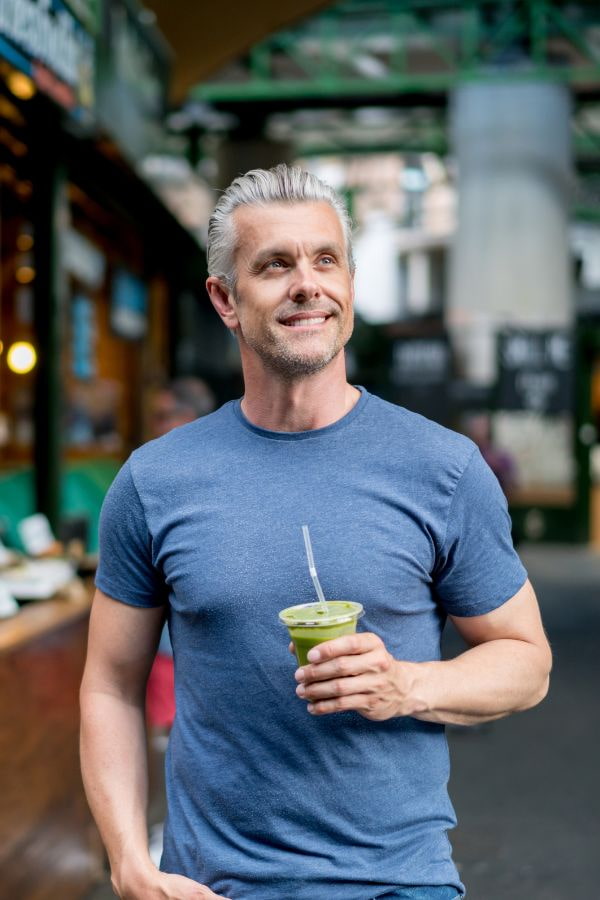 image of a man smiling while walking down a street and holding a cup of green juice